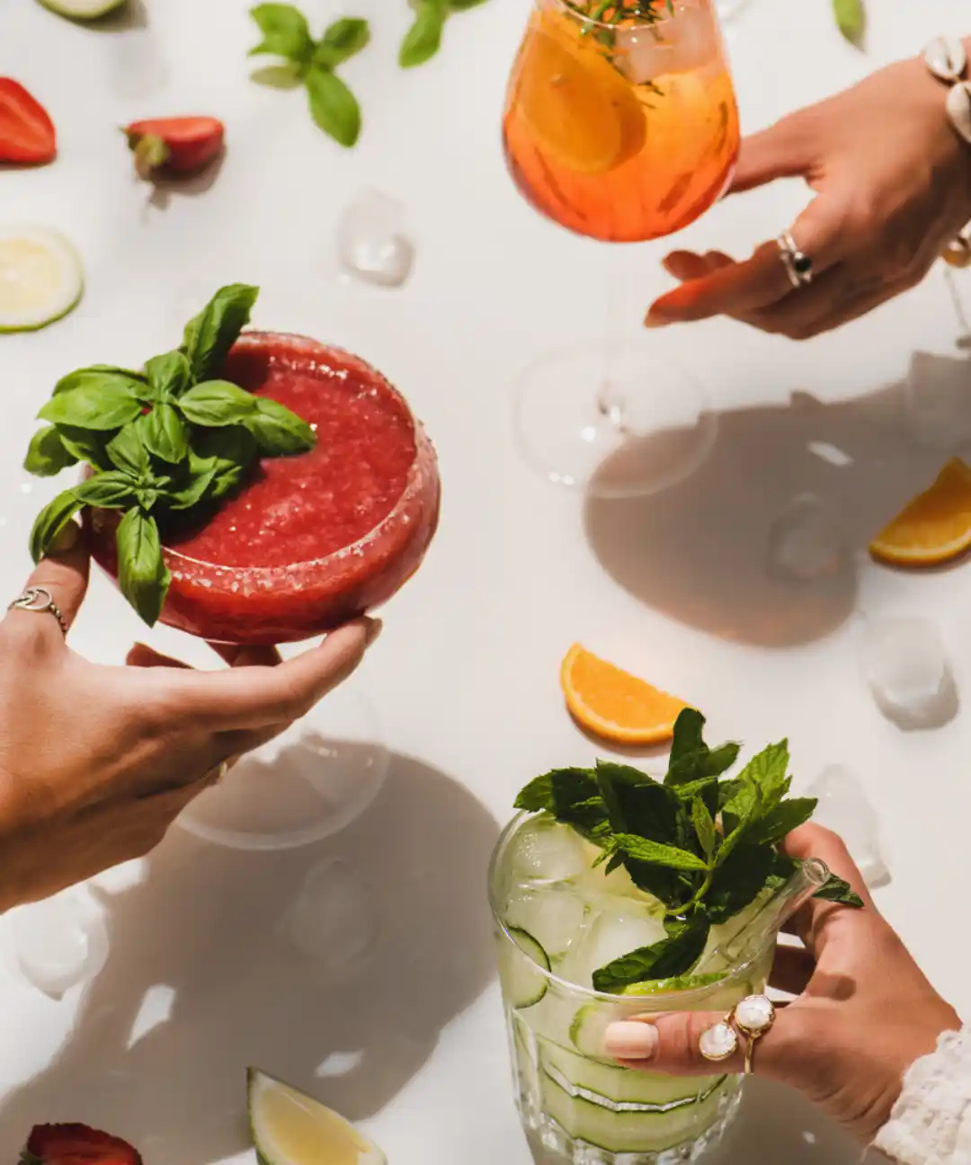 Three colorful non-alcoholic drinks with fresh herbs, citrus, and berries held by hand—perfect for healthy, feel-good recipes.
